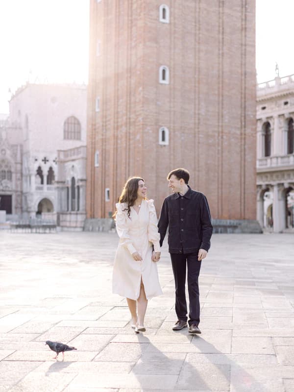 A couple walks hand in hand across St. Mark’s Square in Venice, with the Campanile and surrounding historic buildings behind them