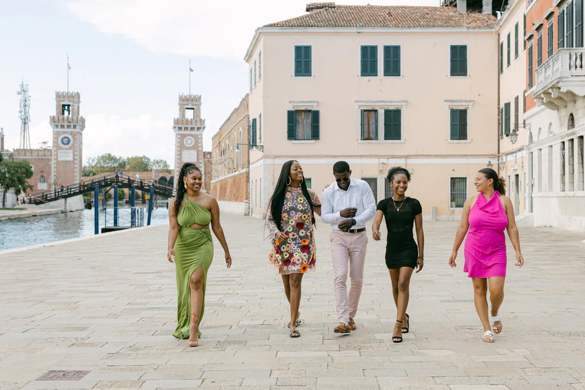 Five friends walking along a Venetian waterfront promenade, with historic towers and canal-side buildings behind them