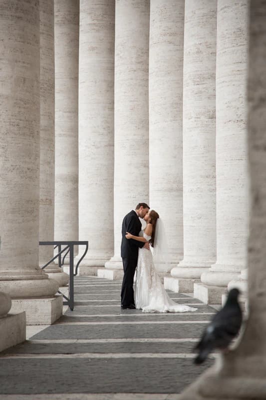 Bride and groom kiss between towering stone columns on the steps of a classical Italian building, with a pigeon in the foreground