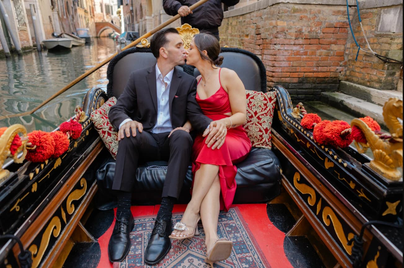 A newlywed couple kisses while seated in an ornate gondola on a narrow Venetian canal