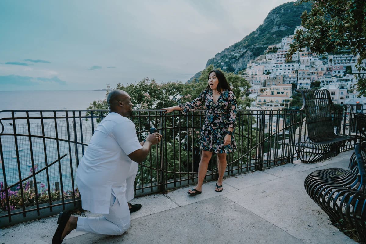 A man kneels before a surprised woman on a seaside terrace overlooking Positano’s cliffside houses on the Amalfi Coast