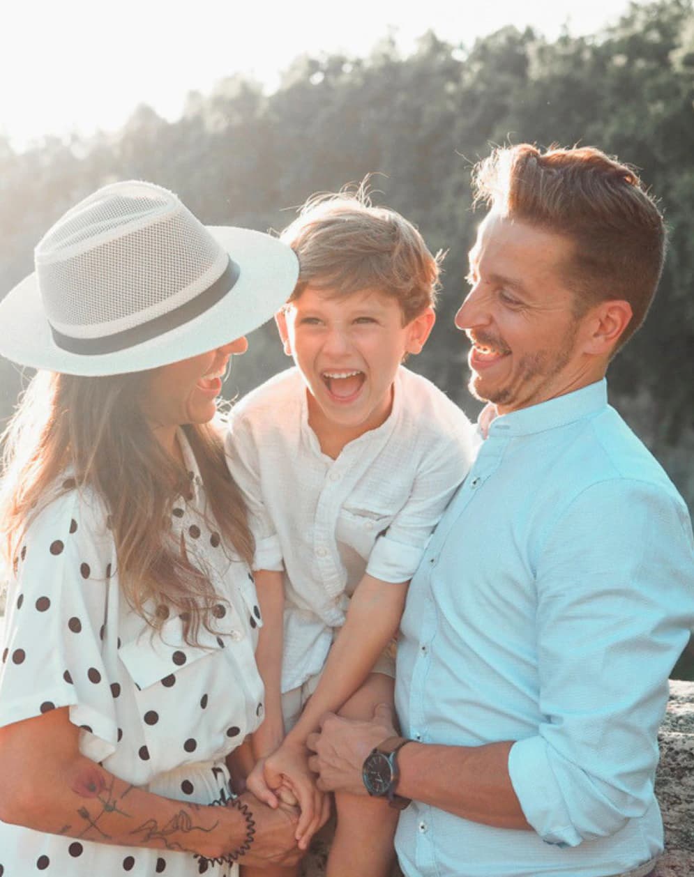 Family smiling together outdoors in warm sunlight, with a mother in a white hat, laughing son, and father holding hands beside a wooded setting