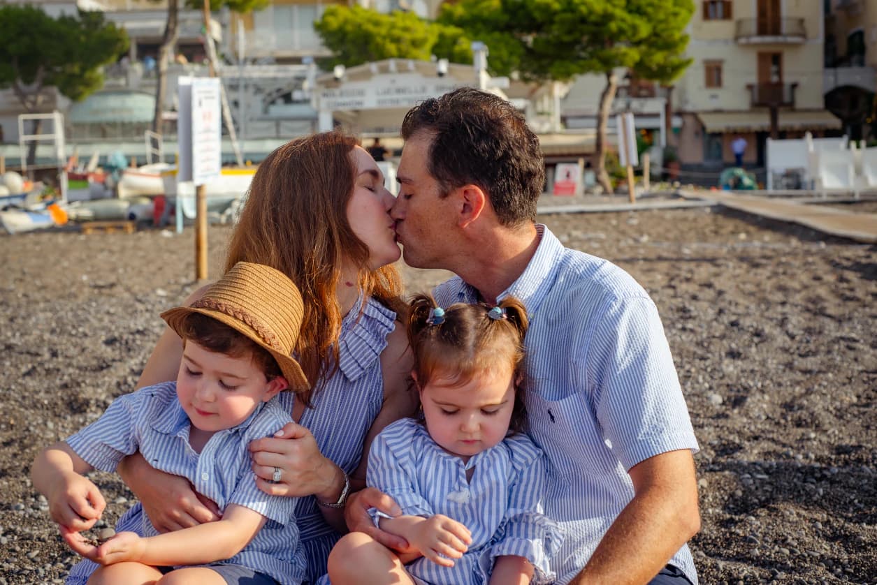 Parents kiss while seated with their two young children on a pebbled beach, with seaside buildings and umbrellas behind them