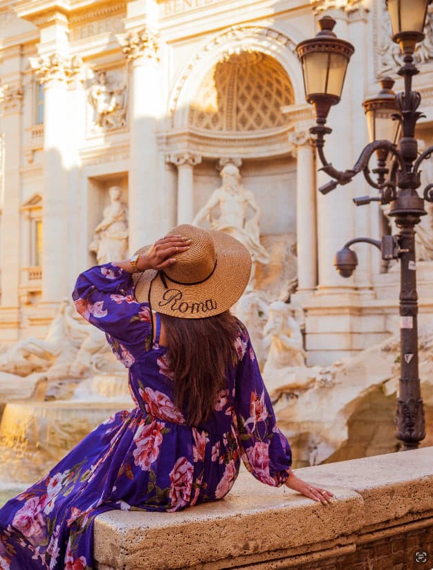 Woman in a purple floral dress and straw hat sitting before Rome’s Trevi Fountain in Italy