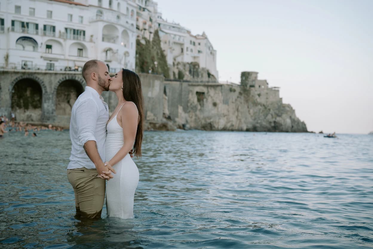 Couple kissing while standing in the sea beneath white cliffside buildings on the Amalfi Coast