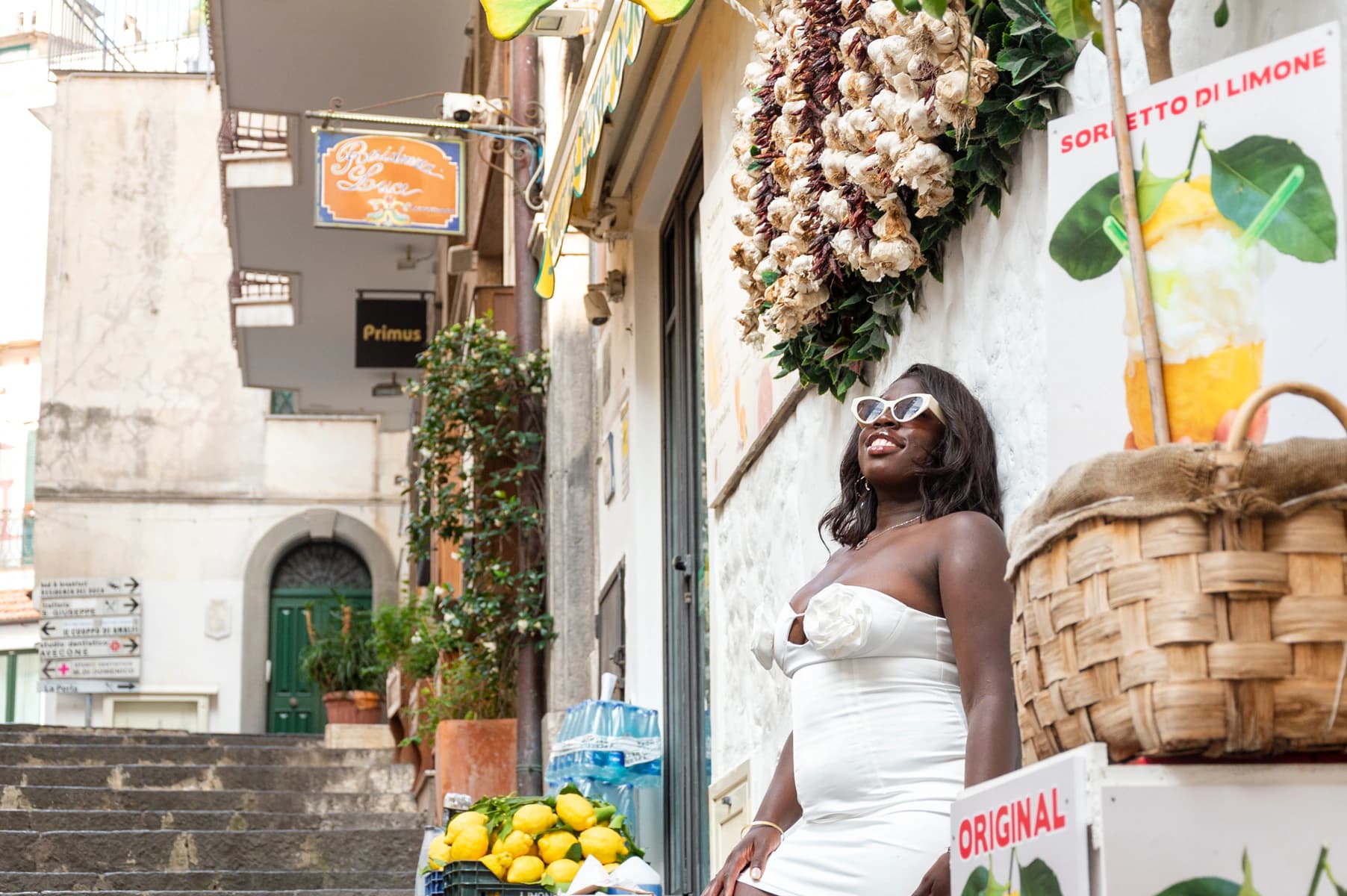 Woman in a white dress standing beside a lemon display on a narrow Italian street lined with shops and stairs